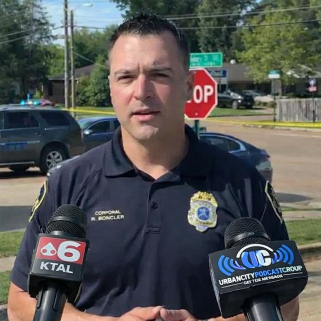 Police officer speaking at a crime scene in Shreveport with two microphones, including Urban City Podcast branding, during investigation of a deadly domestic dispute shooting involving child victims.