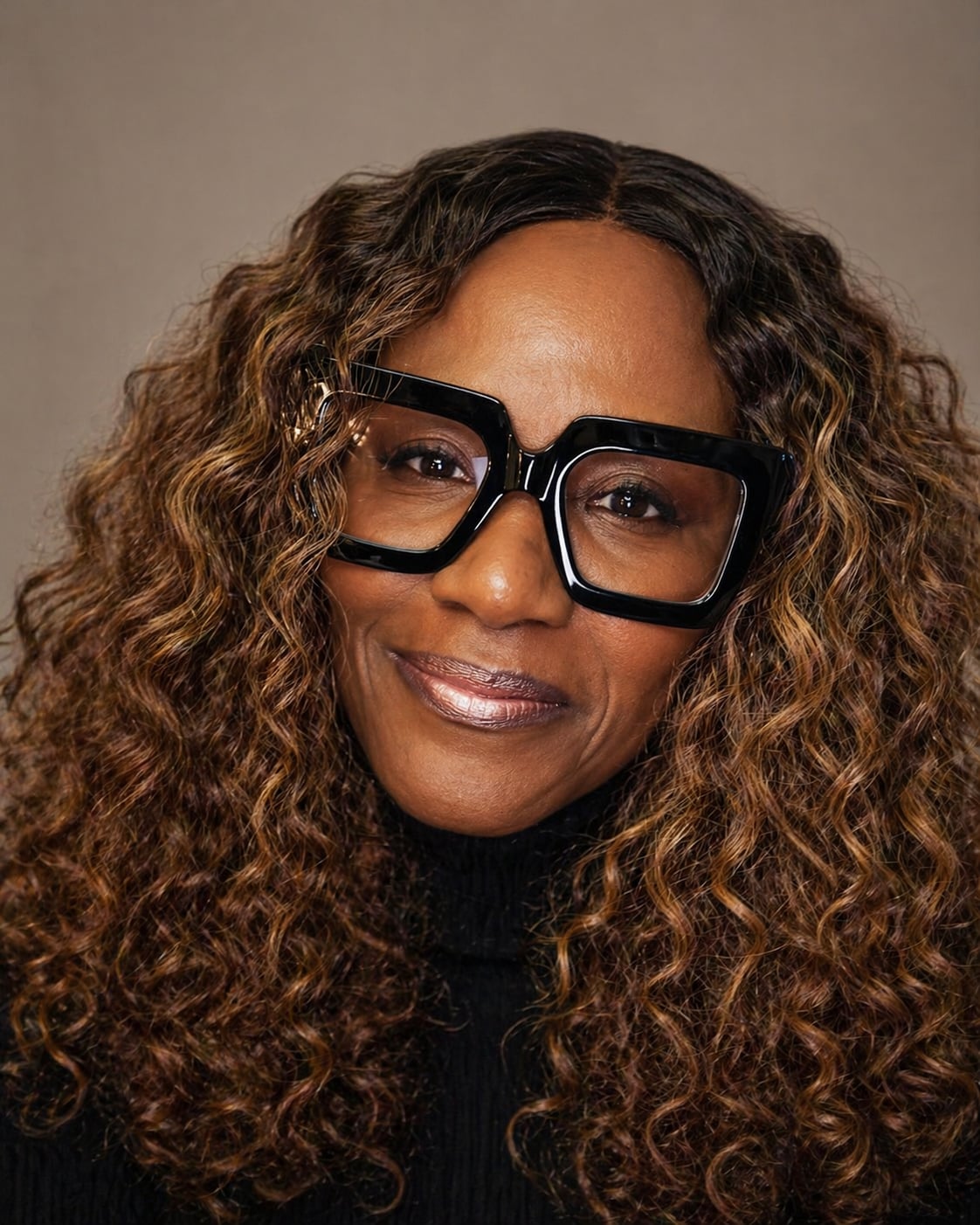 Portrait of author Felicia Brookins wearing oversized black glasses and a black turtleneck, smiling confidently with long curly highlighted hair against a neutral background.