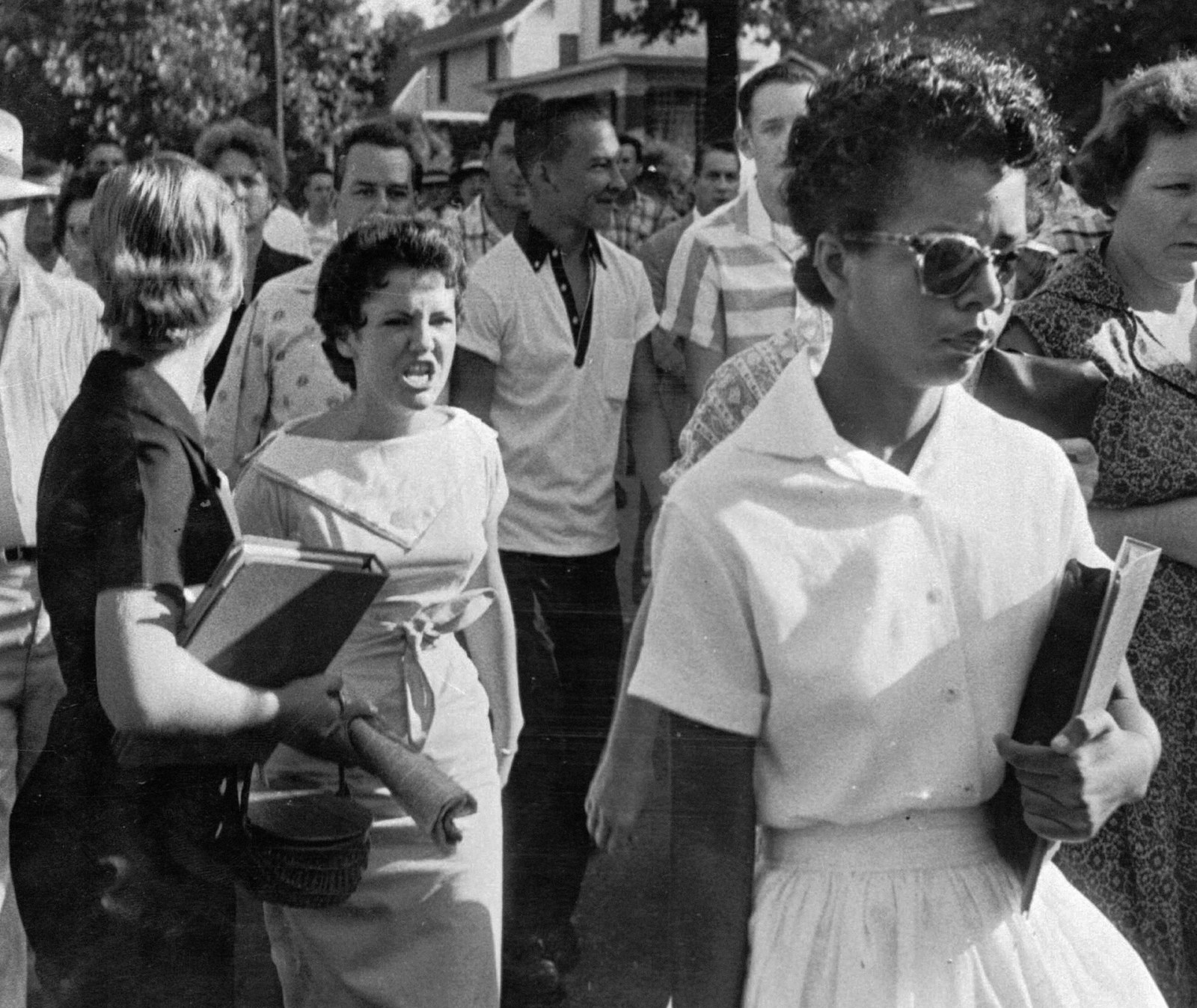 Elizabeth Eckford, wearing sunglasses and a white dress and carrying her school books, walks toward Little Rock Central High School in 1957 while a hostile crowd shouts behind her