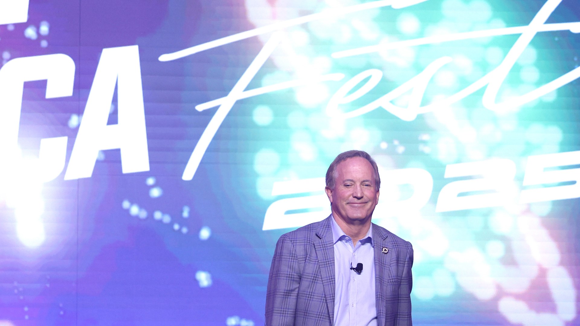 Texas Attorney General Ken Paxton smiling on a stage in a plaid jacket and light blue shirt