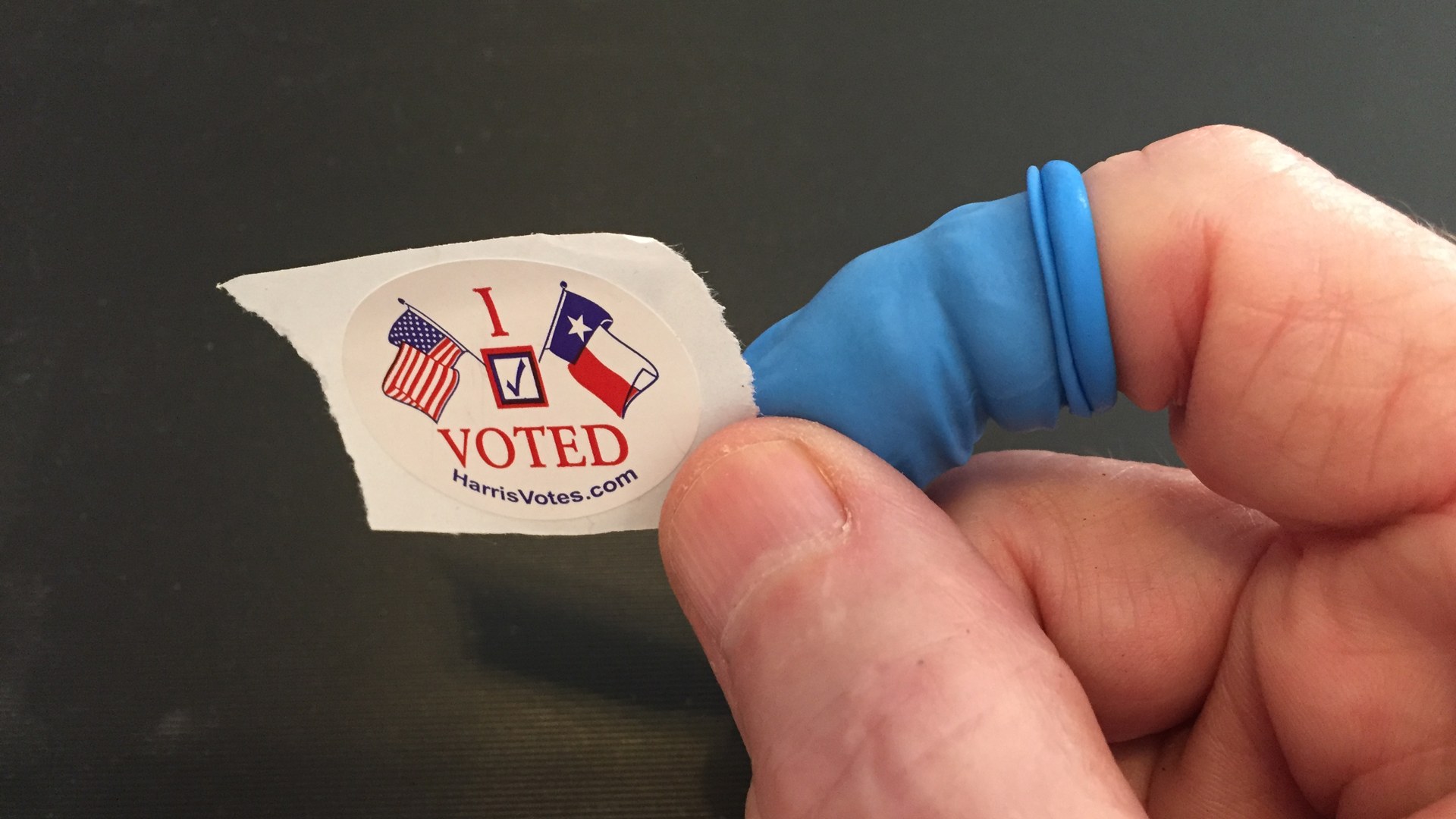 A hand holds a Harris County I Voted sticker with United States and Texas flags on it
