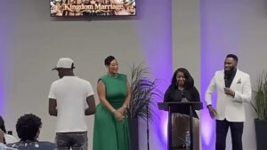 Prophet Israel Ford holds a microphone on stage beside Pastor Lydia Ford at a podium and Minister Shakayla Ford in a green dress during the announcement at LifeWorks Church