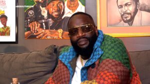 Rick Ross seated during an interview wearing a colorful designer jacket and gold framed glasses