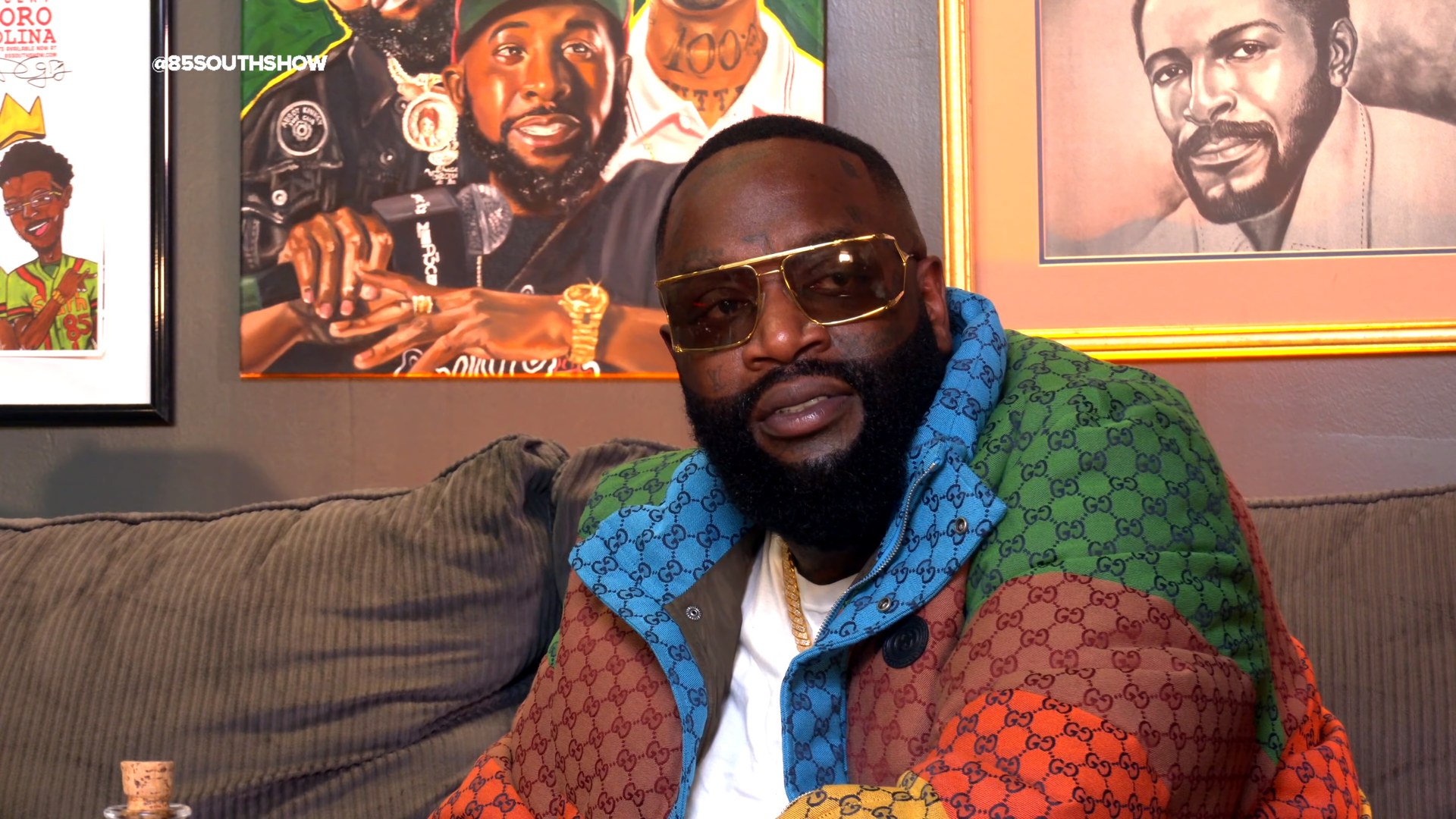 Rick Ross seated during an interview wearing a colorful designer jacket and gold framed glasses