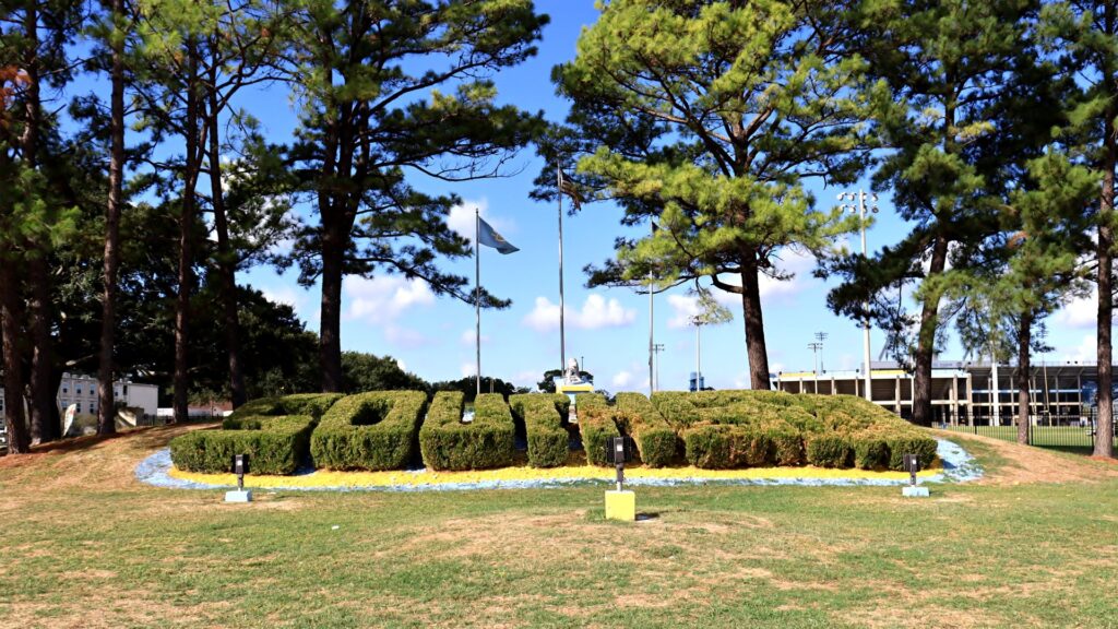 The word Southern spelled out in trimmed hedges on a grassy hill on the Southern University campus in Baton Rouge, with pine trees, flags and the stadium behind it