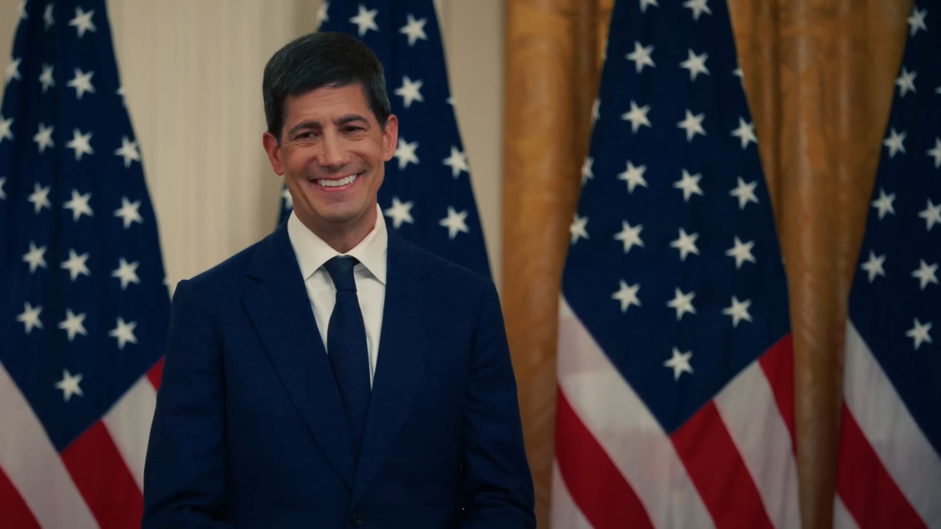Federal Reserve Chair Kevin Warsh smiling in a navy suit in front of United States flags