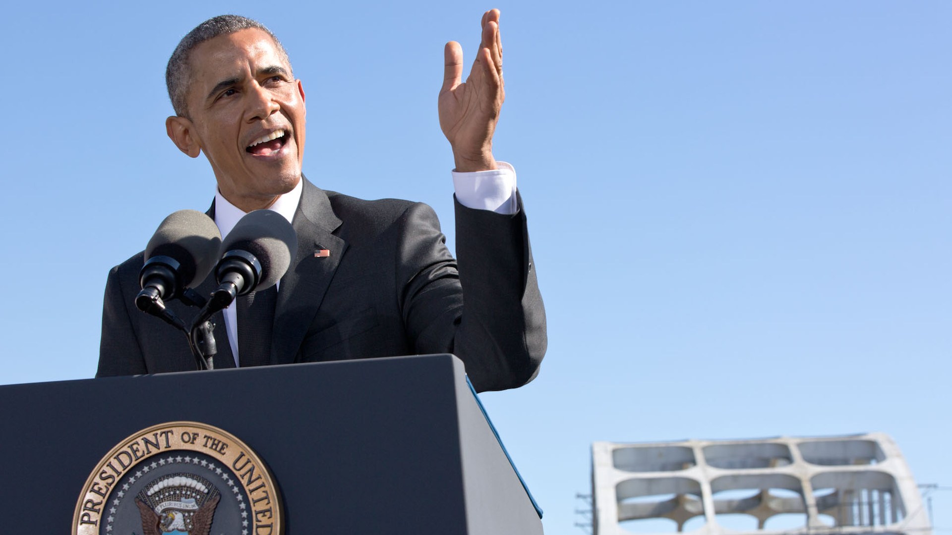 Barack Obama speaking at a podium with the Edmund Pettus Bridge behind him