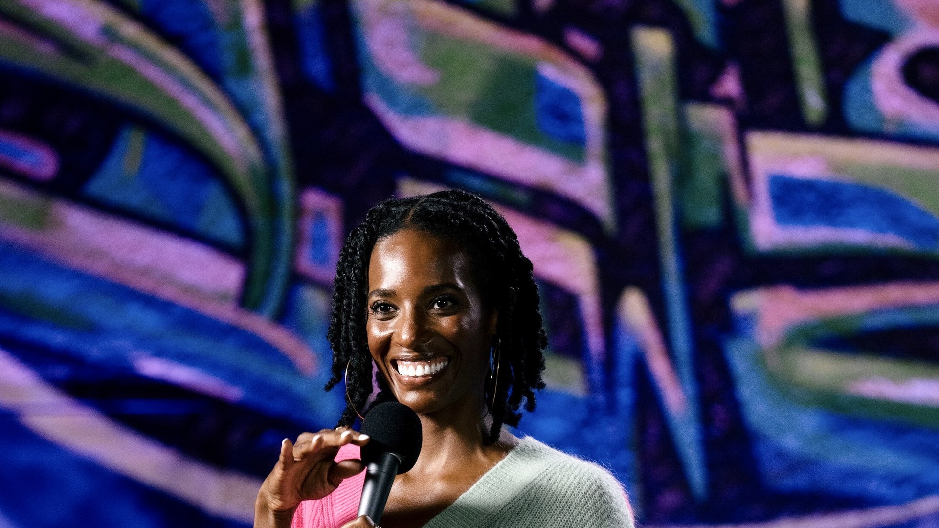 Zainab Johnson smiles on stage in a colorful sweater holding a microphone