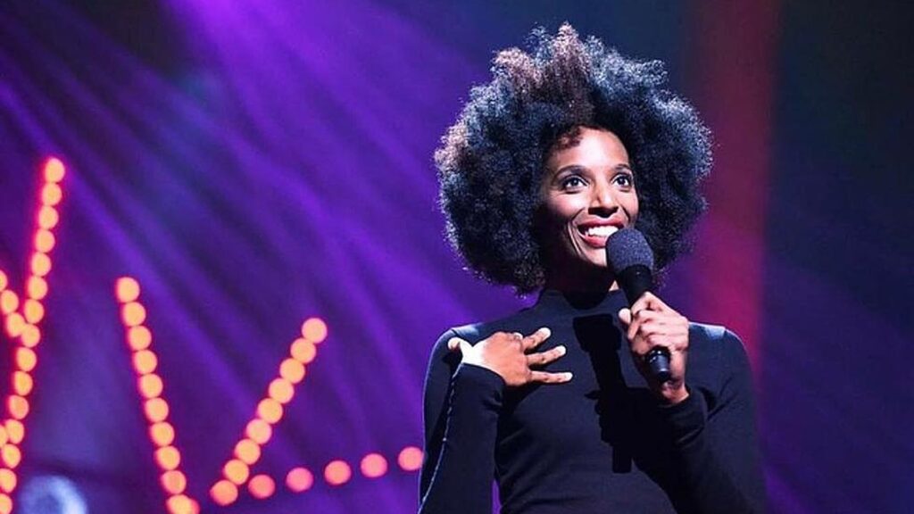 Zainab Johnson performs stand up on stage holding a microphone under purple lights