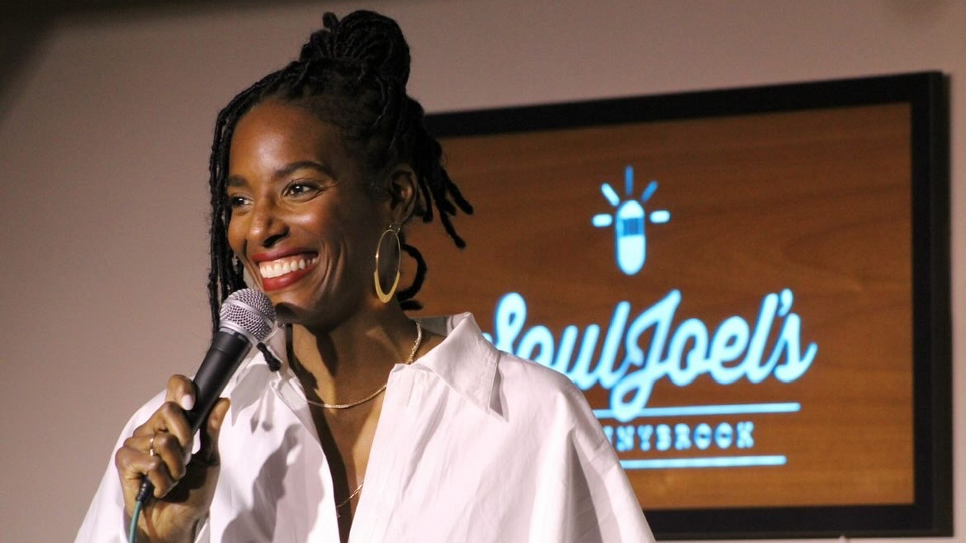 Zainab Johnson laughs while performing stand up in a white shirt holding a microphone